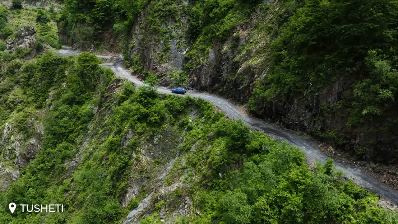 Narrow cliffside gravel road with a blue 4x4 vehicle on a hairpin turn, steep drop and dense green vegetation, Tusheti Abano Pass