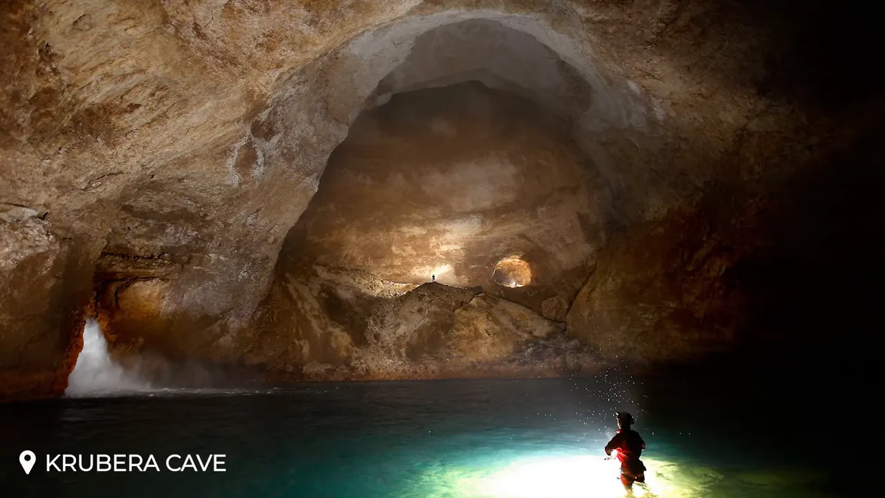 Vast Krubera cave chamber with a caver in illuminated water and small figures on a distant ridge, showing dramatic scale